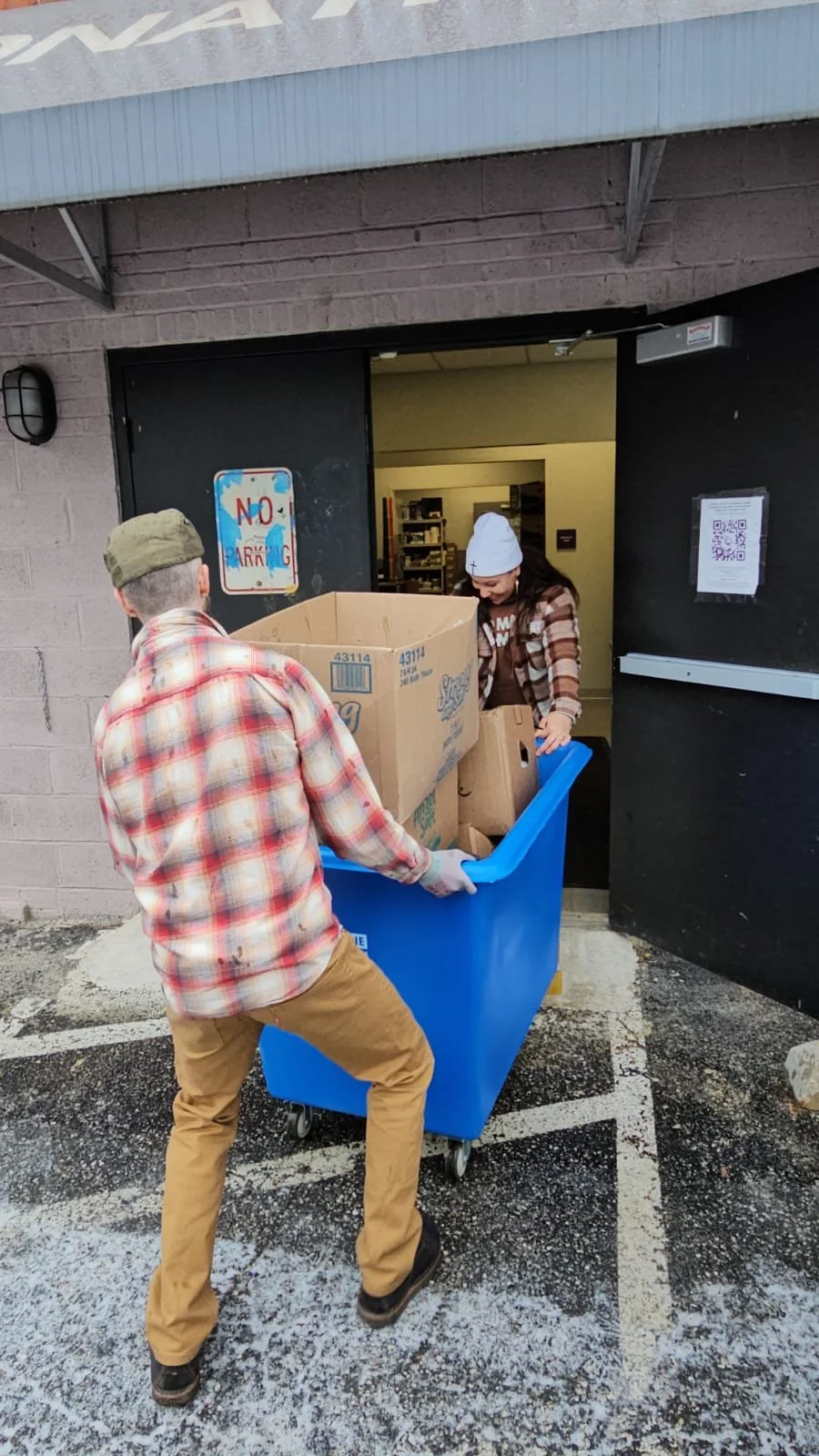 Two people are moving a blue trash bin filled with cardboard boxes outside a building. One person is wearing a beige cap and plaid shirt, and the other is wearing a white beanie and a plaid jacket.