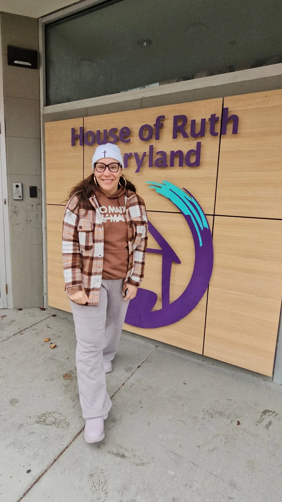 A woman standing in front of a sign that reads "House of Ruth Maryland." She is smiling, wearing glasses, a white beanie, earrings, a brown t-shirt, a plaid jacket, and light-colored sweatpants, outdoors on a sidewalk.