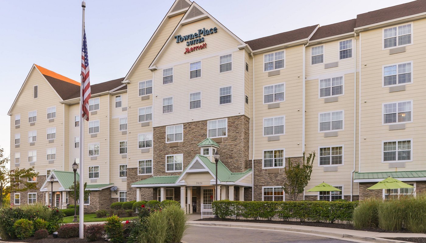 Exterior of a TownePlace Suites by Marriott hotel building with multiple stories, light yellow siding, green roof accents, and a landscaped front area with bushes and outdoor seating, including umbrellas.