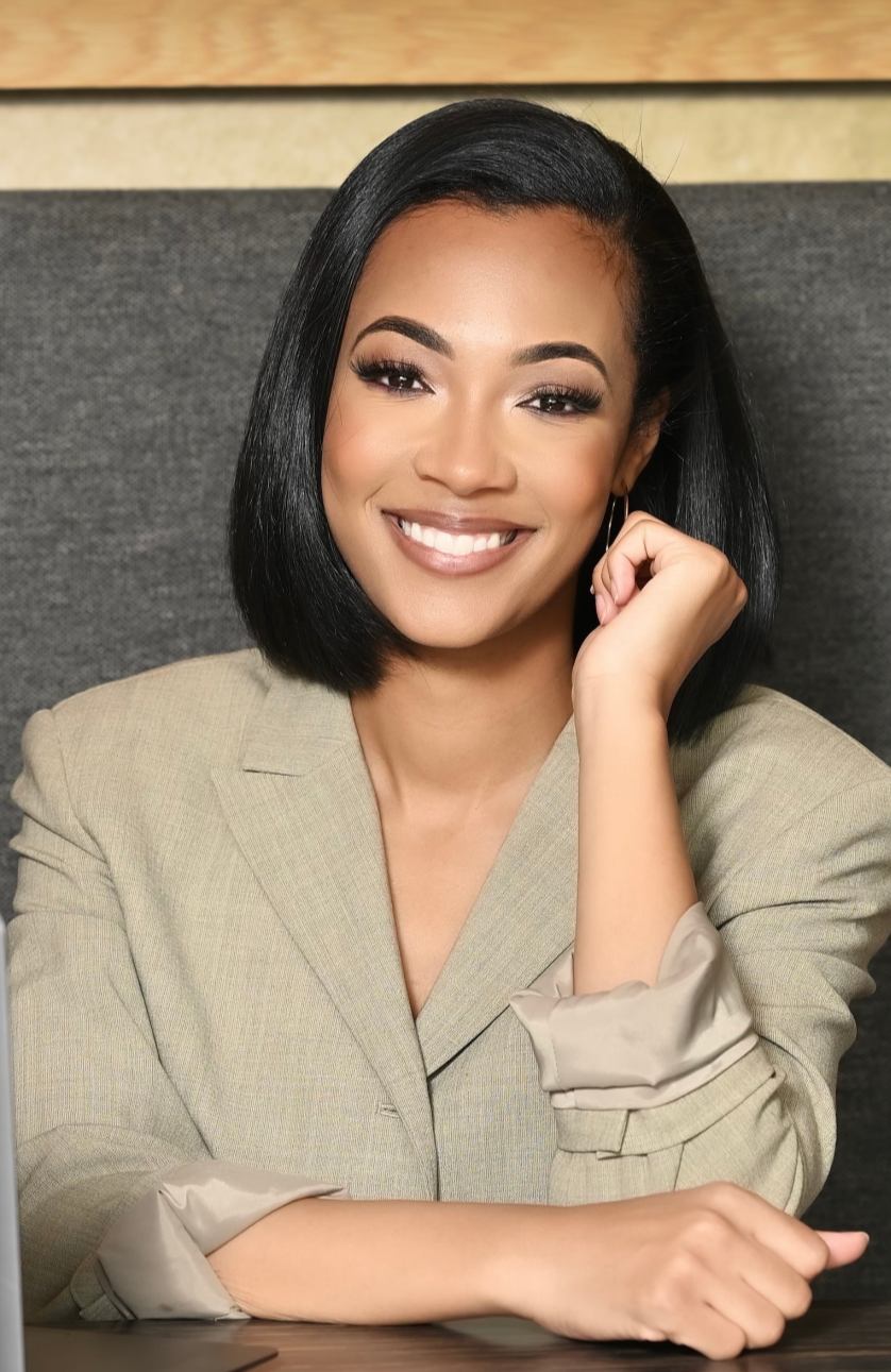 Smiling woman with black hair, wearing a beige blazer, sitting at a desk.