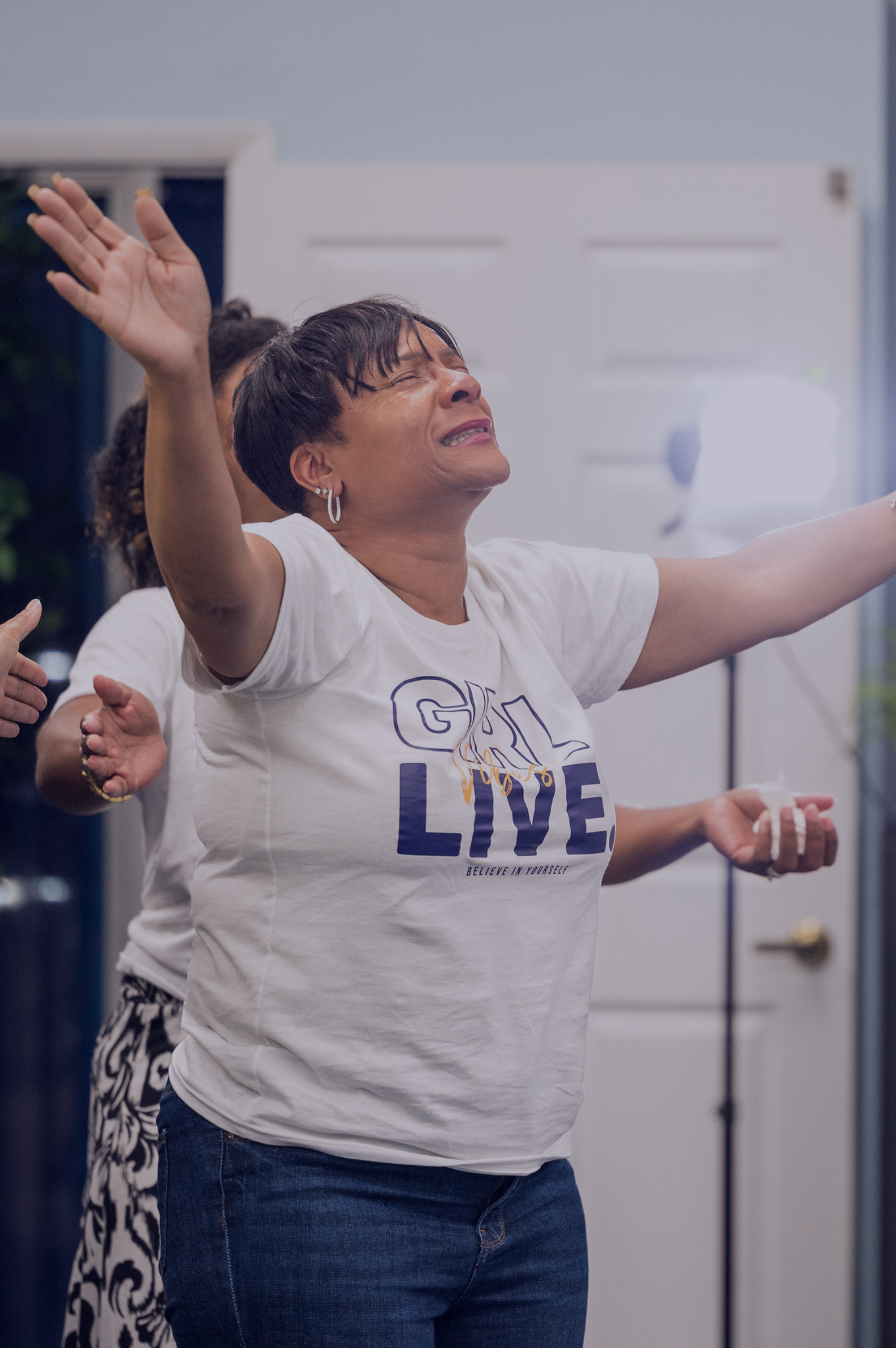 A woman with short dark hair and earrings, wearing a white T-shirt that says 'GIRL LIVE' and 'BELIEVE IN YOURSELF,' has her arms raised with a joyful expression and eyes closed in a moment of celebration or worship.