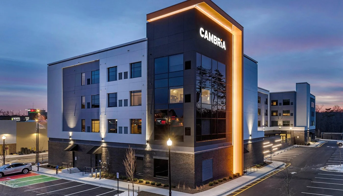 Exterior view of a modern hotel named 'Cambria' during evening with lit-up signage and parking lot