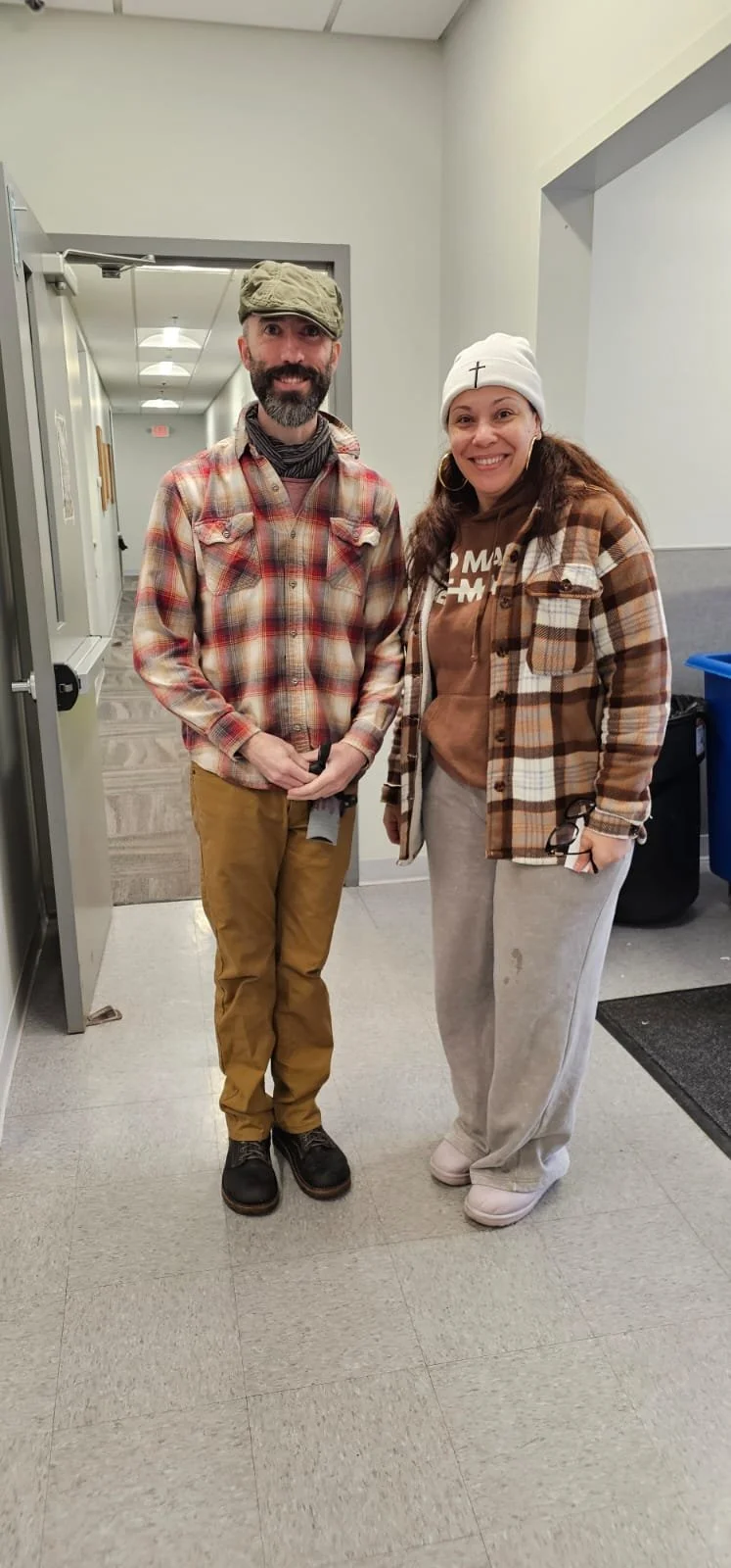 A man and woman standing indoors, both smiling for the photo. They are dressed in casual, fall-appropriate clothing, including flannel shirts and beanies.