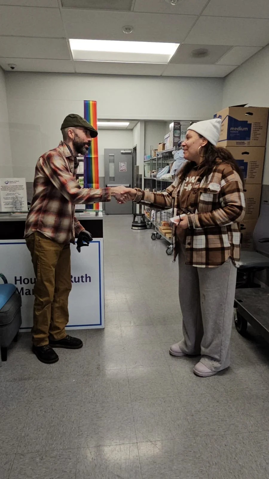 A man and woman shaking hands in a room with shelves and boxes, both wearing plaid shirts.
