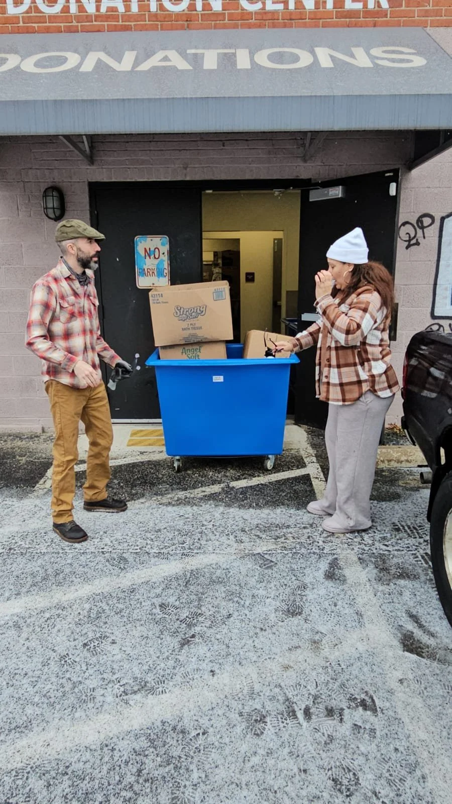 Two people stand outside a donation center next to a blue recycling bin filled with boxes. The person on the left is a man wearing a green cap, plaid shirt, and tan pants. The person on the right is a woman wearing a white beanie, plaid shirt, and gr