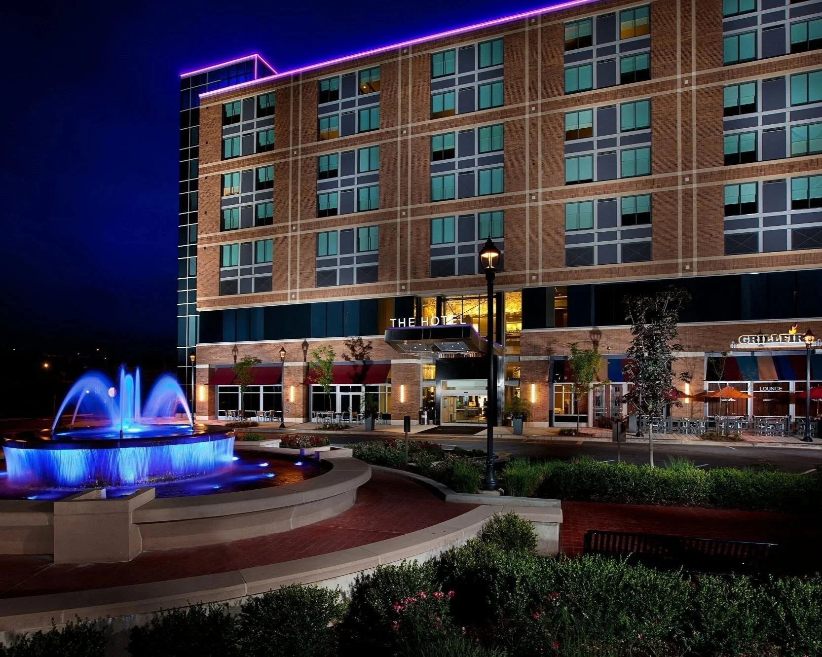Night view of a multi-story hotel with illuminated purple neon accents on the roof. The hotel has a brick facade with numerous windows. In front, there is a lit fountain with blue water jets and a seating area with tables, chairs, and umbrellas. The hotel entrance has a sign that reads 'The Hotel', and nearby are signs for a bar or lounge, and outdoor seating areas.