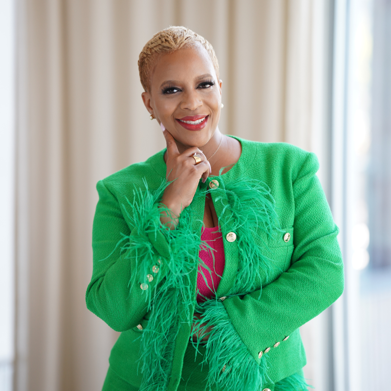 A woman wearing a vibrant green jacket with feathered detailing and gold buttons, smiling and posing with her hand on her chin in front of a neutral background.