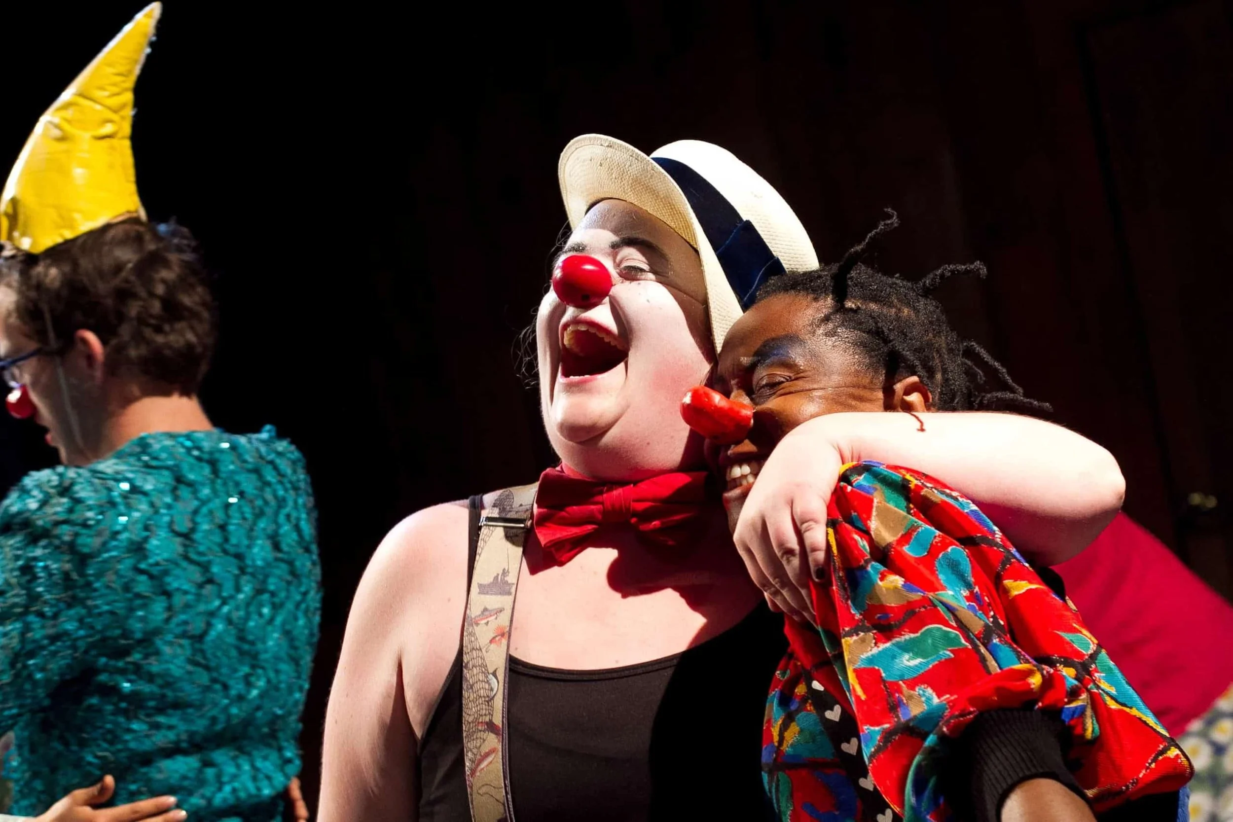 Group of people celebrating, with two individuals dressed as clowns; one wearing a red nose and hat, the other with an orange nose and a colorful jacket, sharing a joyful moment.