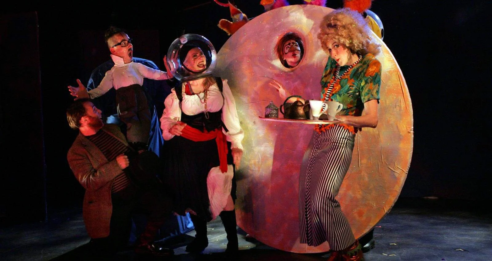 People in colorful costumes performing on stage with a large, round, pink, and orange prop resembling a teapot or a play area. The scene appears theatrical and whimsical.