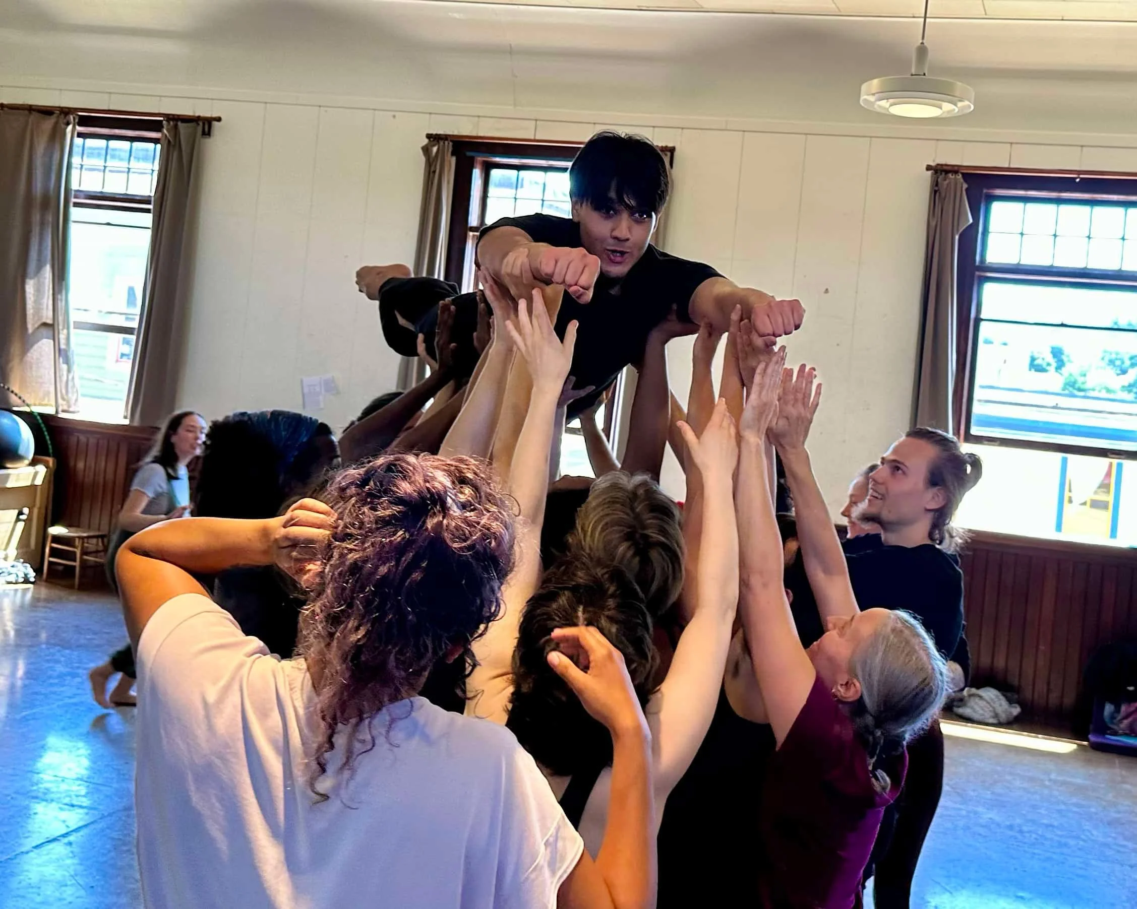 People participating in a dance circle, holding hands and forming a tower inside a room with large windows and natural light.