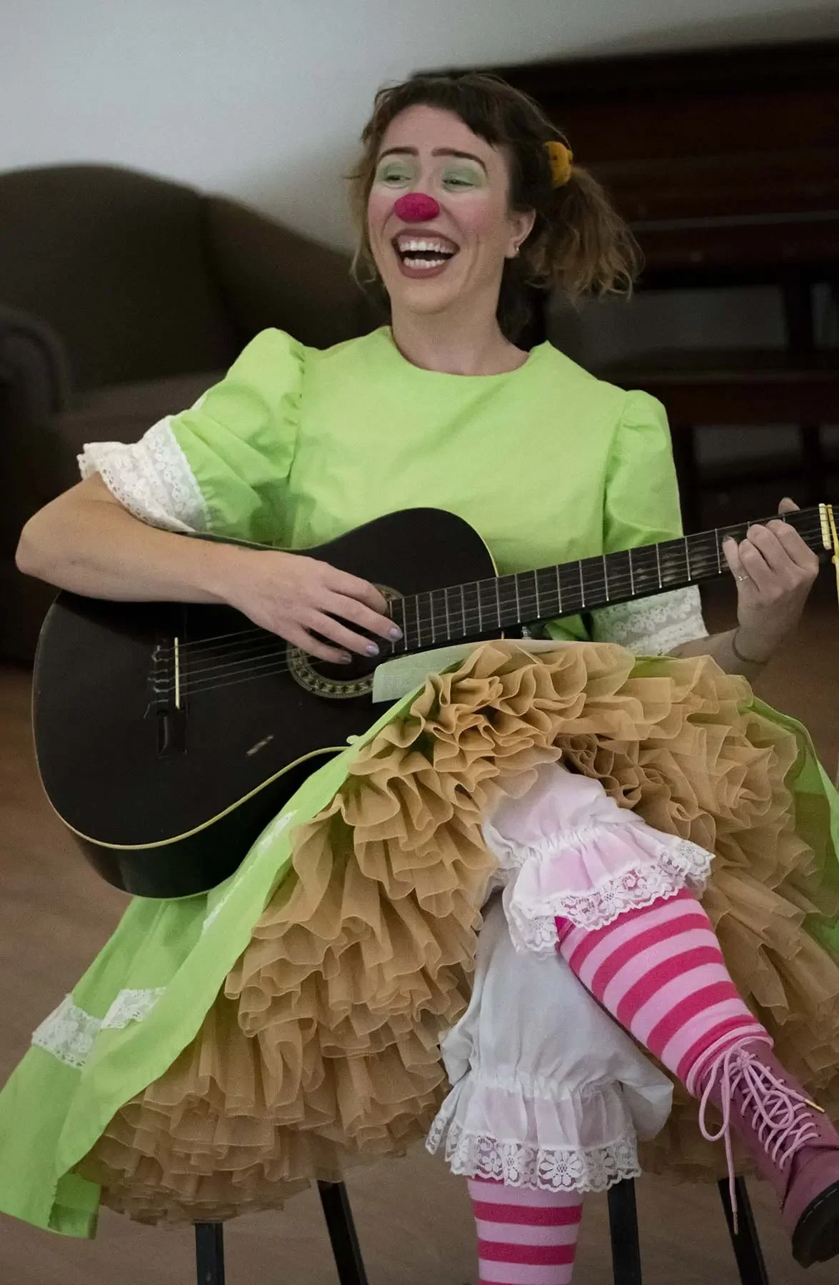 A woman dressed as a clown, wearing a green dress with white lace details, a red nose, green makeup around her eyes, and a yellow hair accessory. She is sitting on a chair, playing a black acoustic guitar, with a ruffled, layered skirt and pink and white striped stockings.