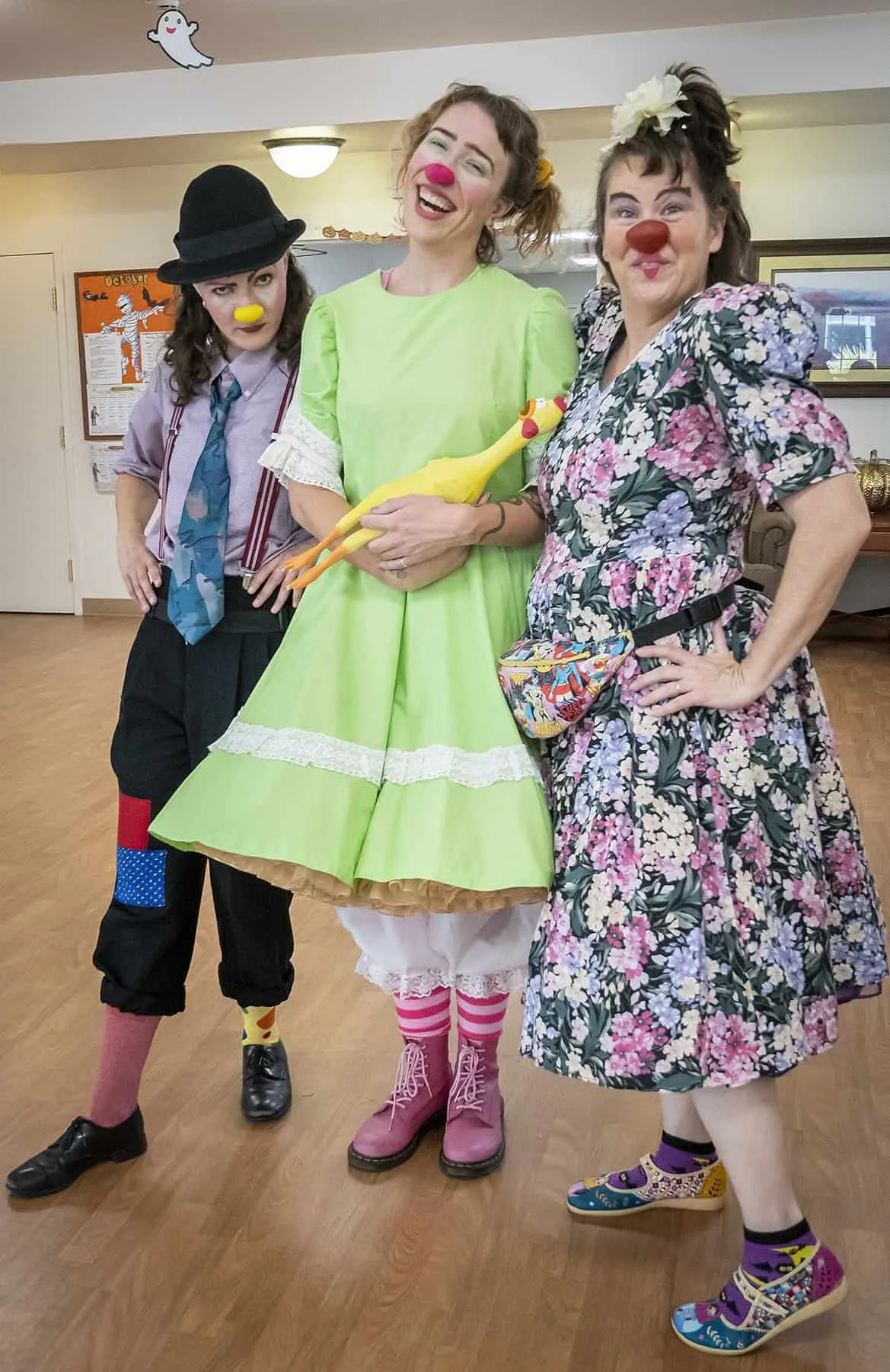 Three women dressed as clowns posing together in a room. They are wearing colorful, playful costumes with clown makeup, including red noses and exaggerated expressions. One woman is holding a rubber chicken.