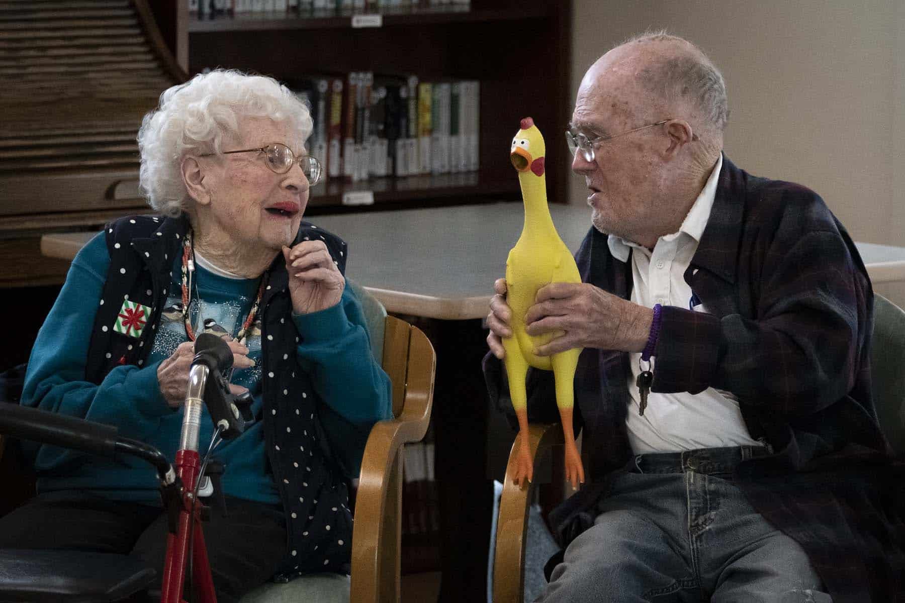 An elderly woman with curly white hair, glasses, and a blue sweater, and an elderly man with glasses and a plaid jacket, sitting at a table. The man is holding a yellow rubber chicken with an animated face, and they are engaging in a conversation indoors, with books on a shelf in the background.