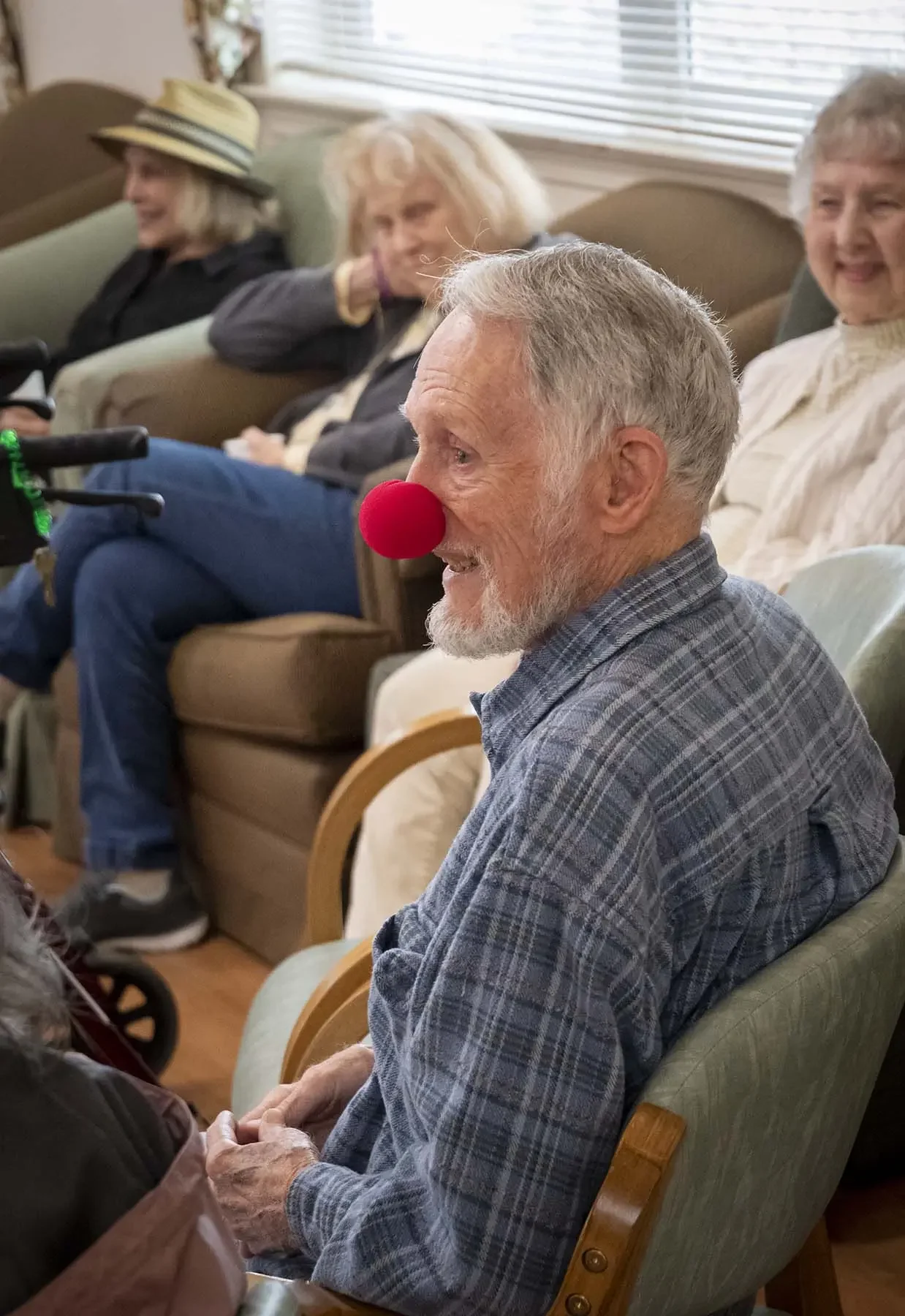 An elderly man with a red clown nose is smiling and holding hands with someone. He has gray hair and a beard, wearing a plaid shirt, sitting in a wooden chair. In the background, three women are seated on couches, smiling and chatting in a cozy room with horizontal blinds on the window.