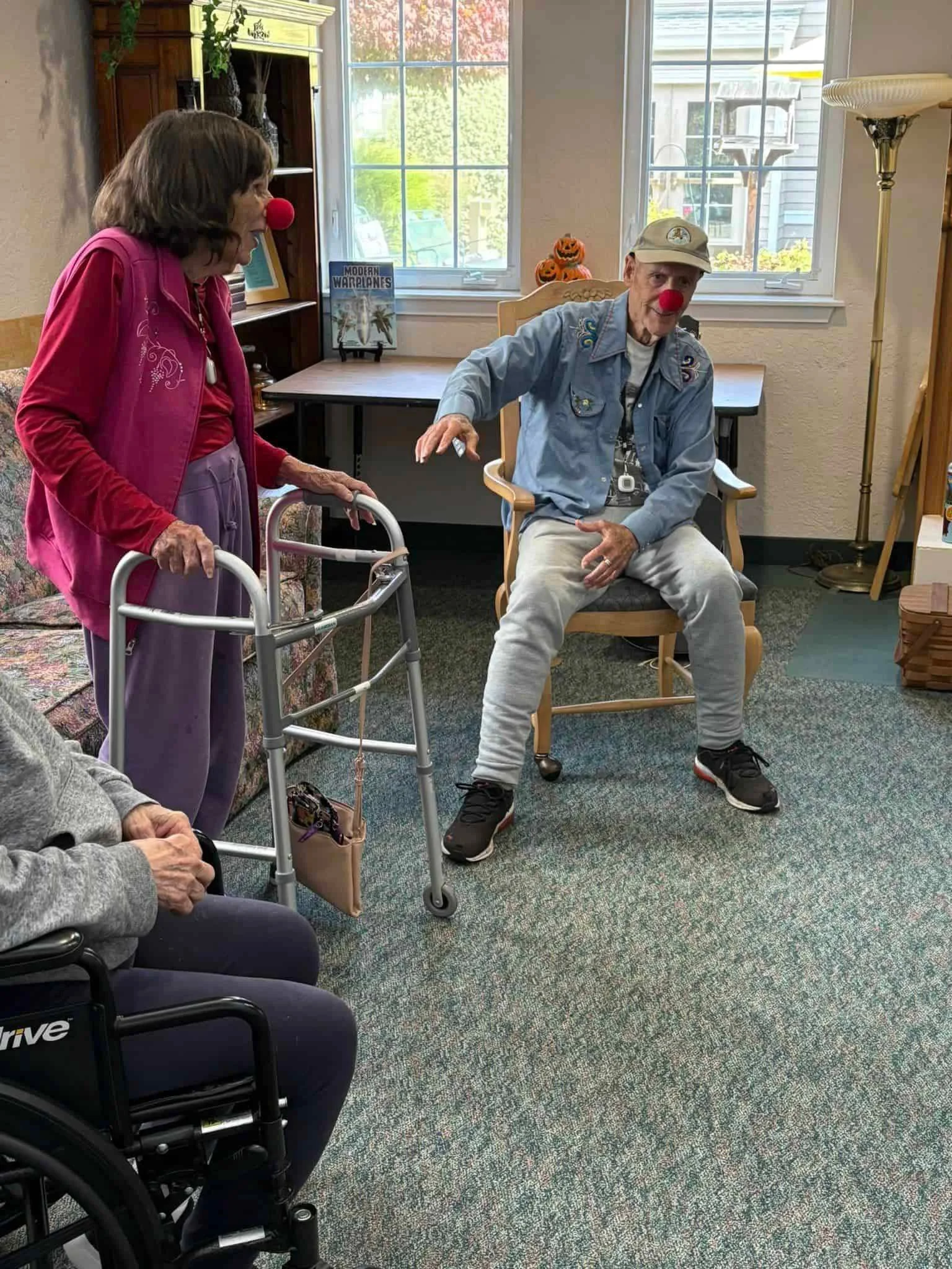 An elderly man with a red clown nose sitting in a wooden rocking chair indoors, gesturing with his right hand, wearing a beige cap and casual clothes, while a woman with a red clown nose and walker stands beside him in a room with large windows, a bookshelf with pumpkin decorations