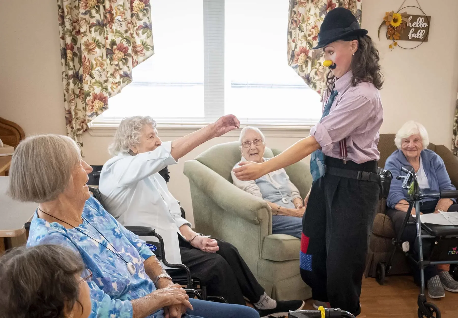 A male clown with a nose and mustache costume entertains senior women in a room with floral curtains and a "Hello Fall" sign on the wall. Some women are in wheelchairs and are smiling or watching the performance.