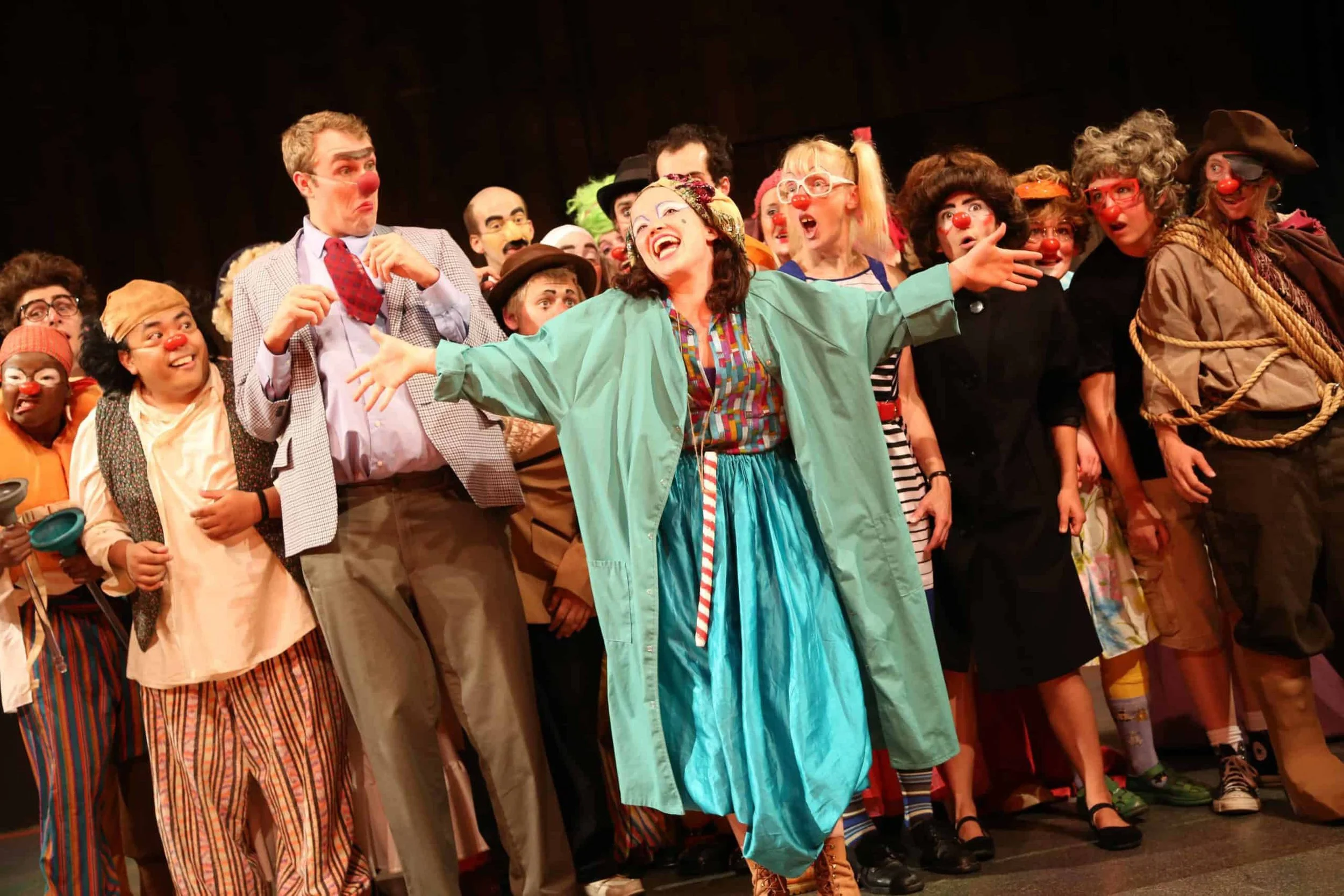 Group of performers in colorful costumes and clown makeup on stage, smiling and engaging with the audience during a theatrical performance.
