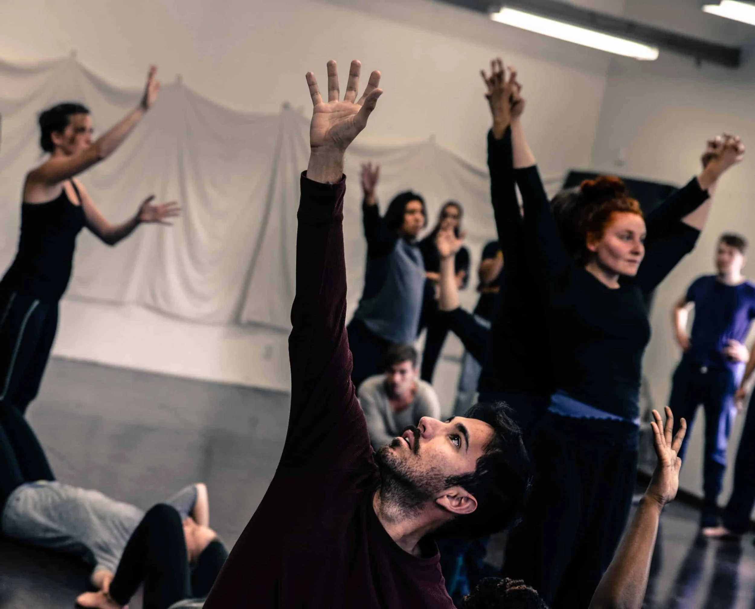 A group of people participating in a yoga or meditation class, with some raising their arms and others in poses, guided by an instructor on the floor.