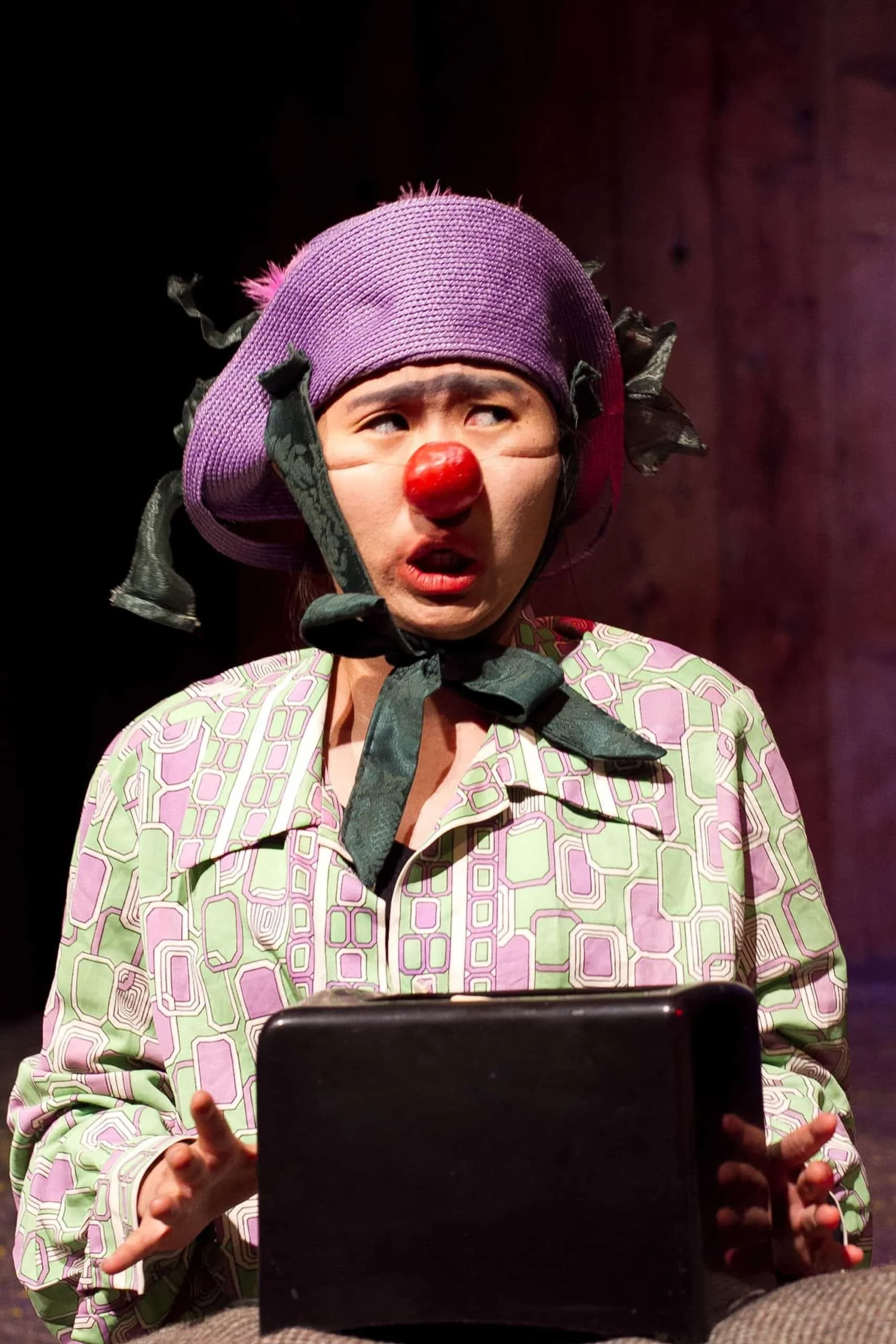 A woman dressed as a clown with a purple hat and red nose looks confused or surprised while holding a black tablet and standing in front of a dark background.