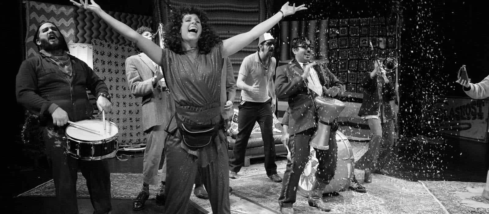 A lively band performing on stage, with a woman in the center dancing with arms raised, surrounded by musicians playing drums, guitar, and trombone, with confetti in the air.