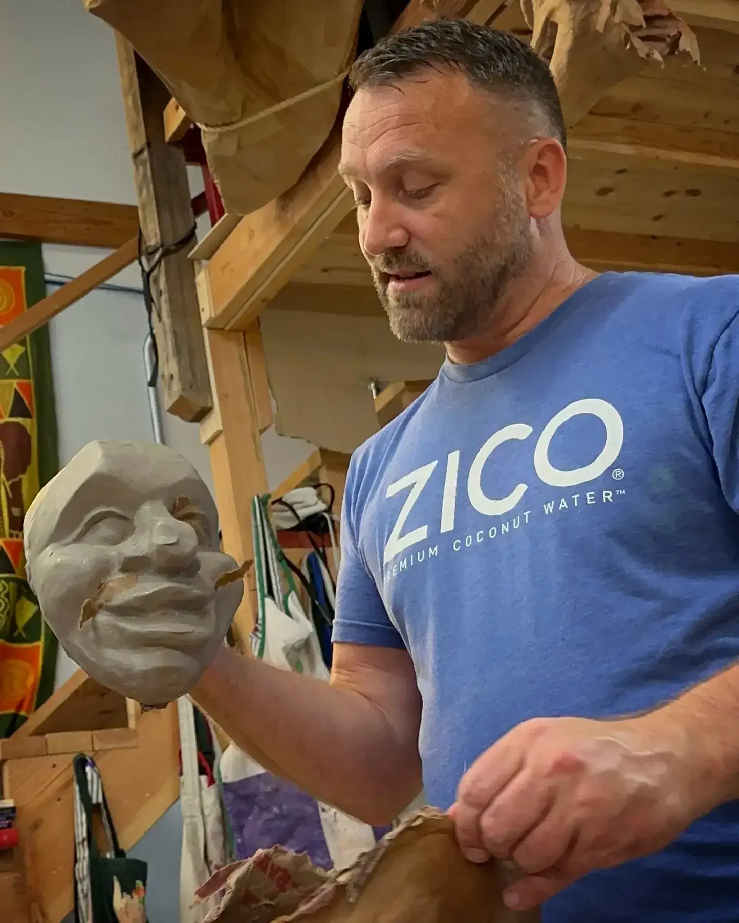 A man is sculpting a detailed face on a clay bust in a woodworking shop. He is wearing a blue Zico t-shirt and has a short beard. The background includes wooden shelves and tools.
