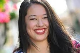 A woman with long dark hair smiling outdoors with blurred flowers in the background.