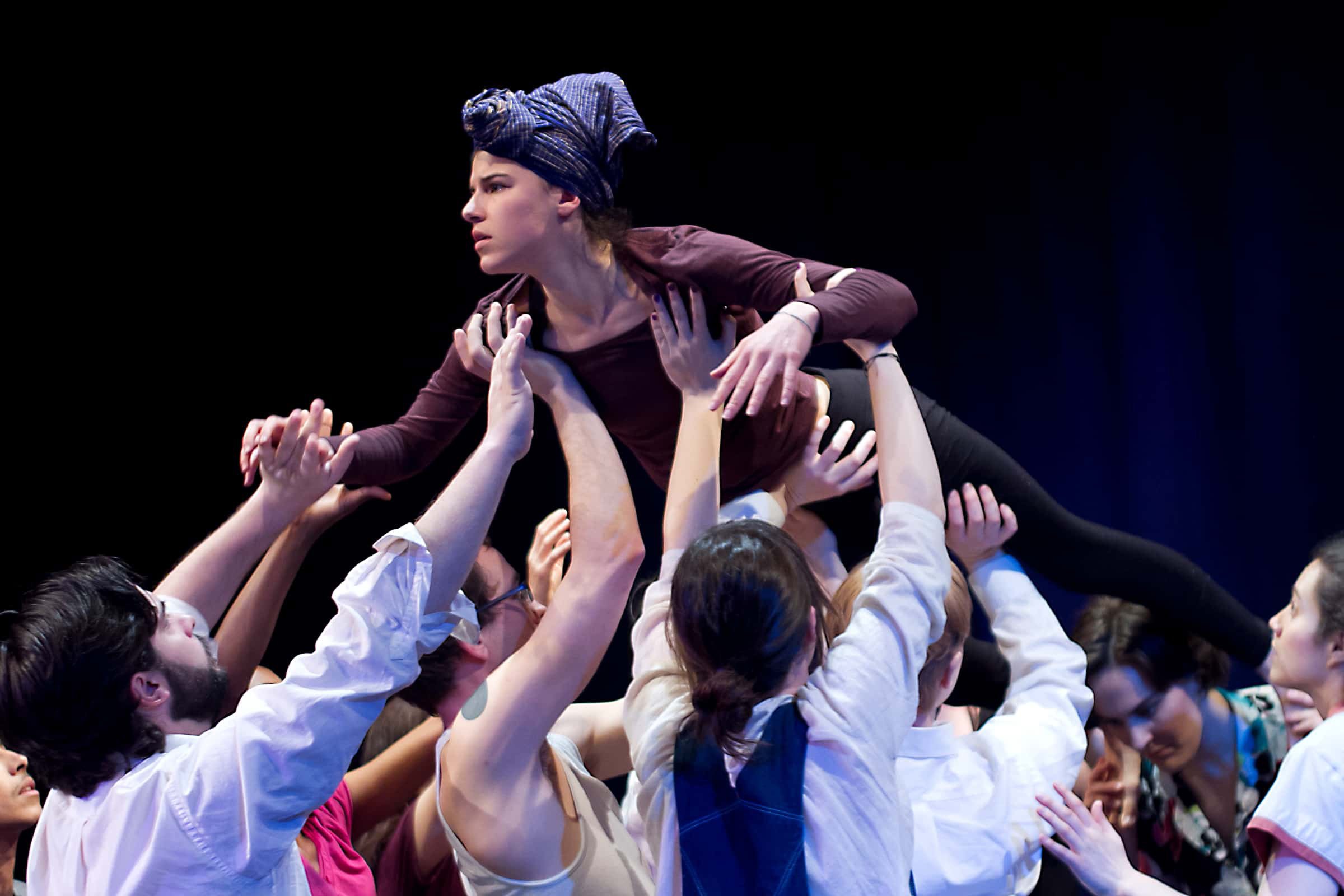 Women and men participating in a theater or performance art, lifting a woman with a blue headscarf. She appears to be in a pose representing concern or focus.