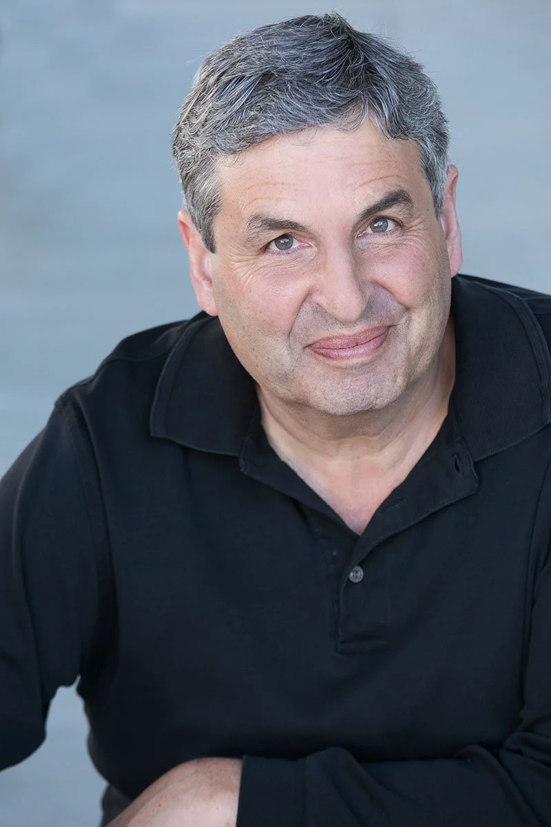 A middle-aged man with short, gray hair and light skin, wearing a black collared shirt, smiling softly at the camera with a plain gray background.