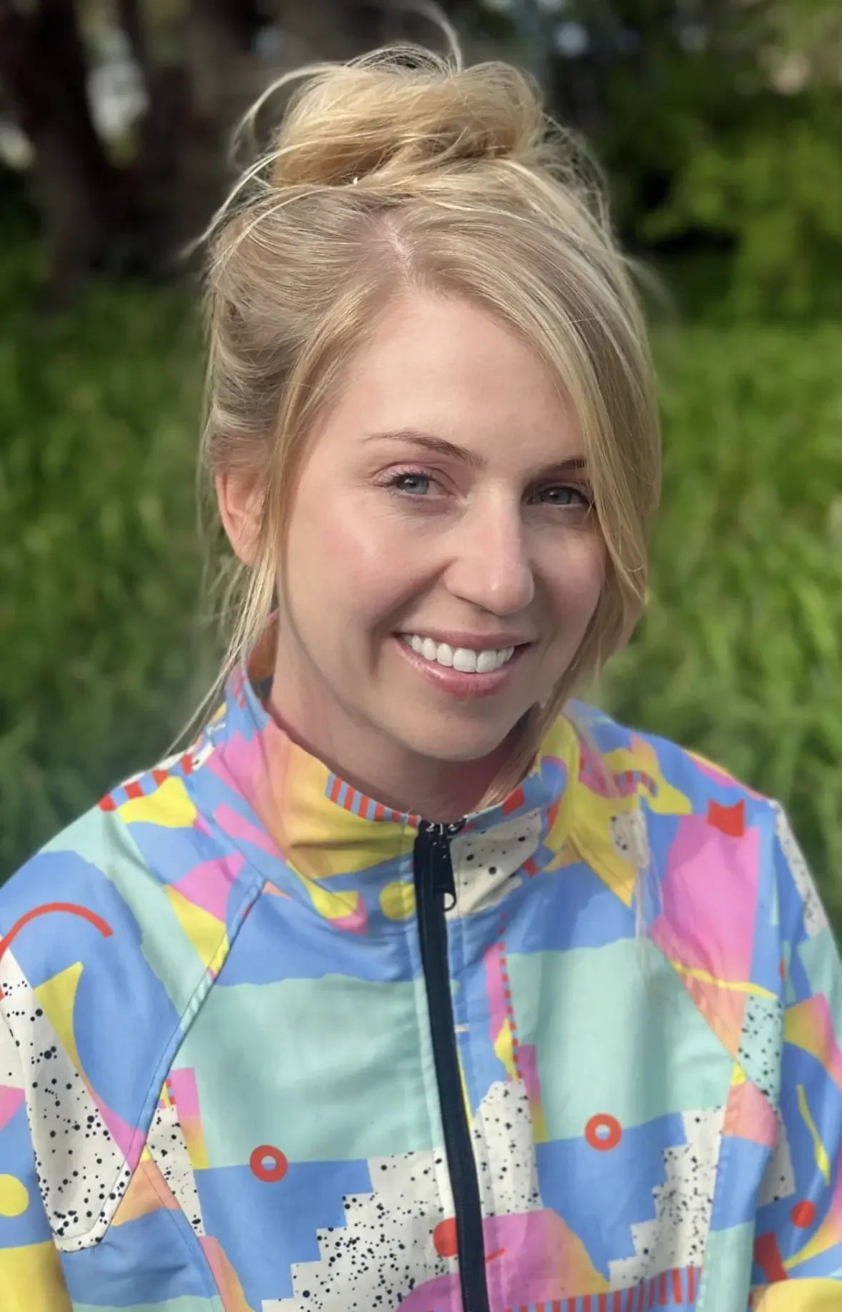 Woman with blonde hair tied up, smiling, wearing a colorful abstract patterned jacket outdoors with green foliage in the background.