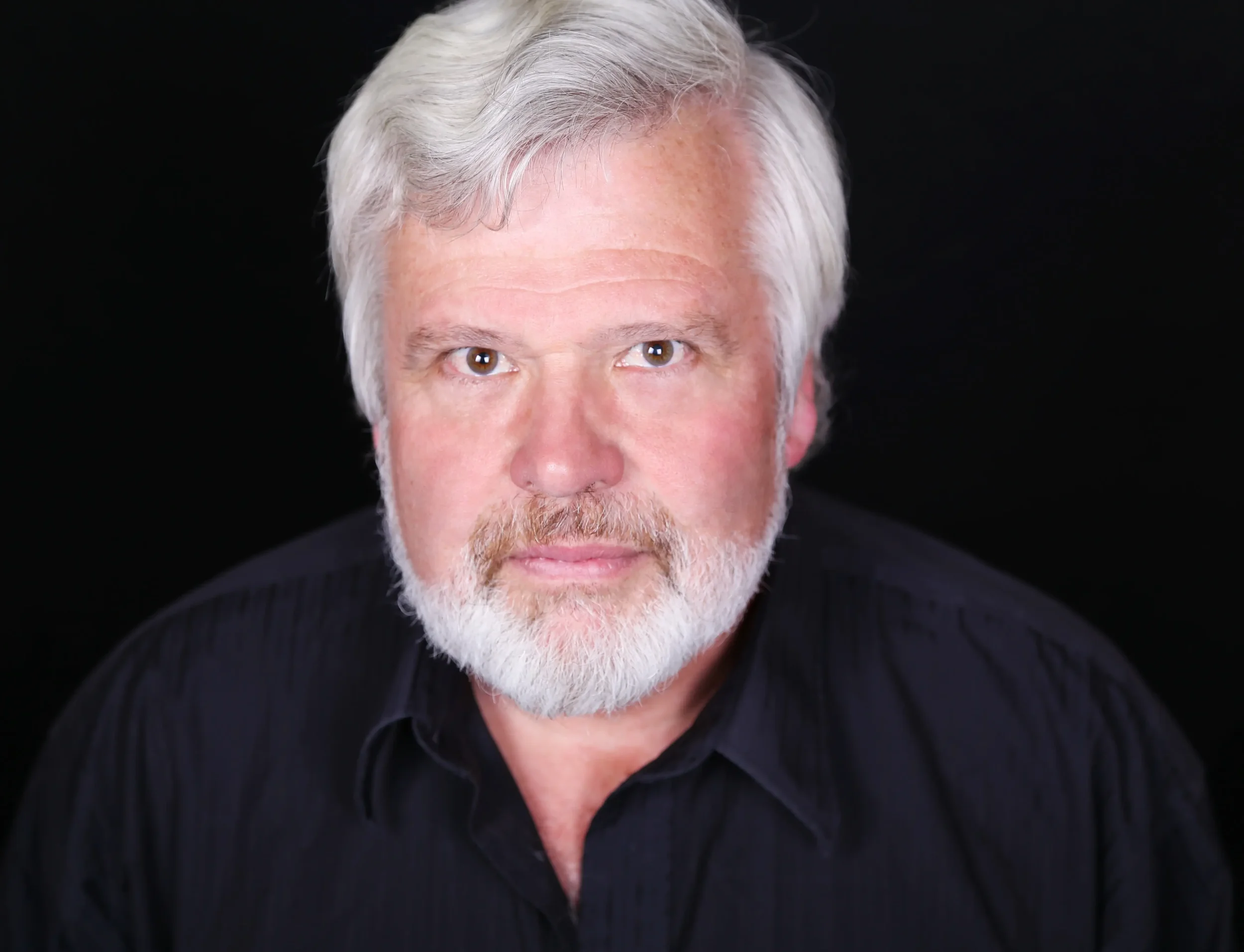 Close-up headshot of an older man with white hair, beard, and mustache, wearing a black shirt, against a black background.