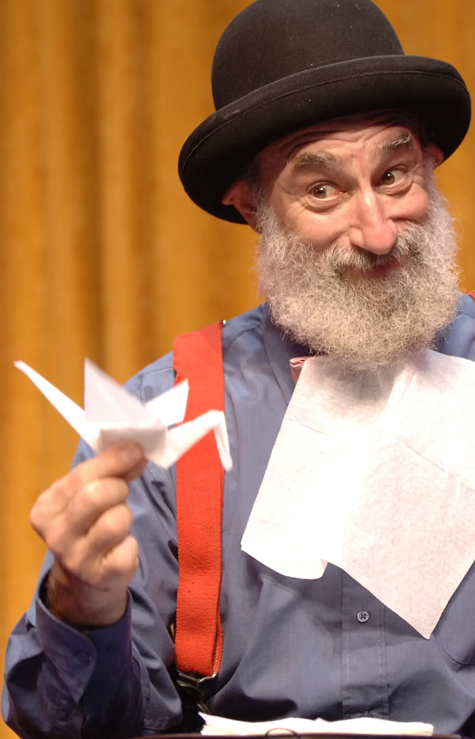 A man with a grey beard, wearing a black top hat, blue shirt, and red suspenders, smiling and holding a paper airplane in front of a yellow curtain.
