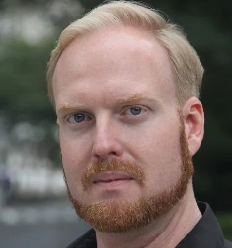 A close-up of a man with blonde hair and a beard, looking directly at the camera outdoors.