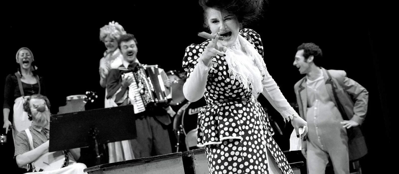 A group of performers on stage, with one woman in the foreground wearing a polka dot outfit and holding a cigar, while others are laughing and playing instruments, in black and white.