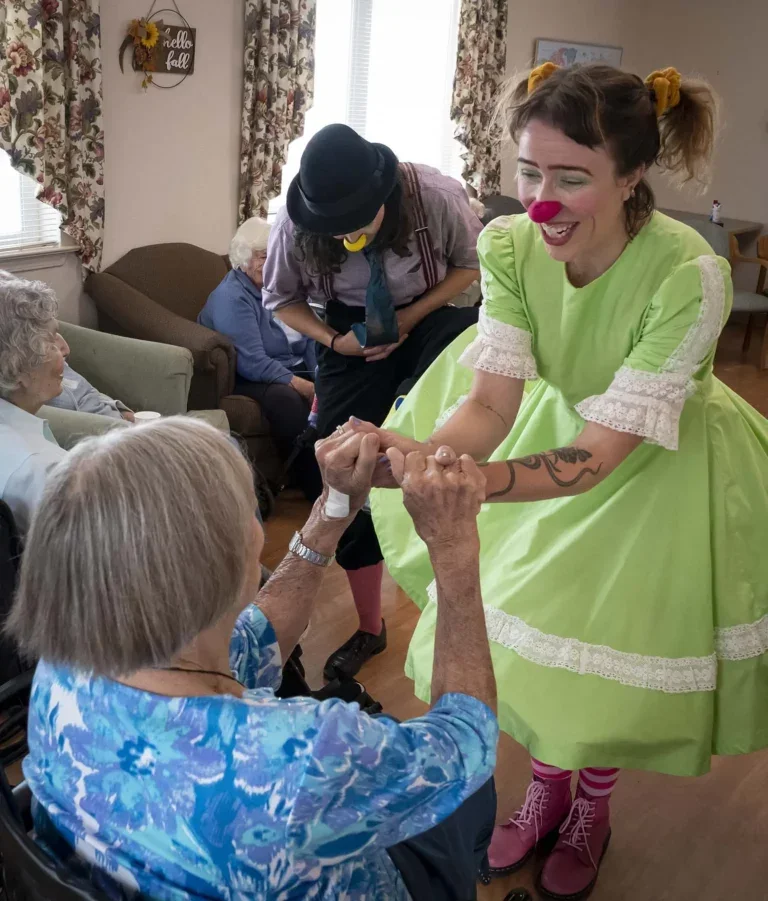 A woman dressed as a clown with green dress, pink shoes, yellow hair ties, and a pink nose, holding hands and smiling at an elderly woman in a blue patterned shirt in a nursing home setting with other elderly residents and staff in the background.