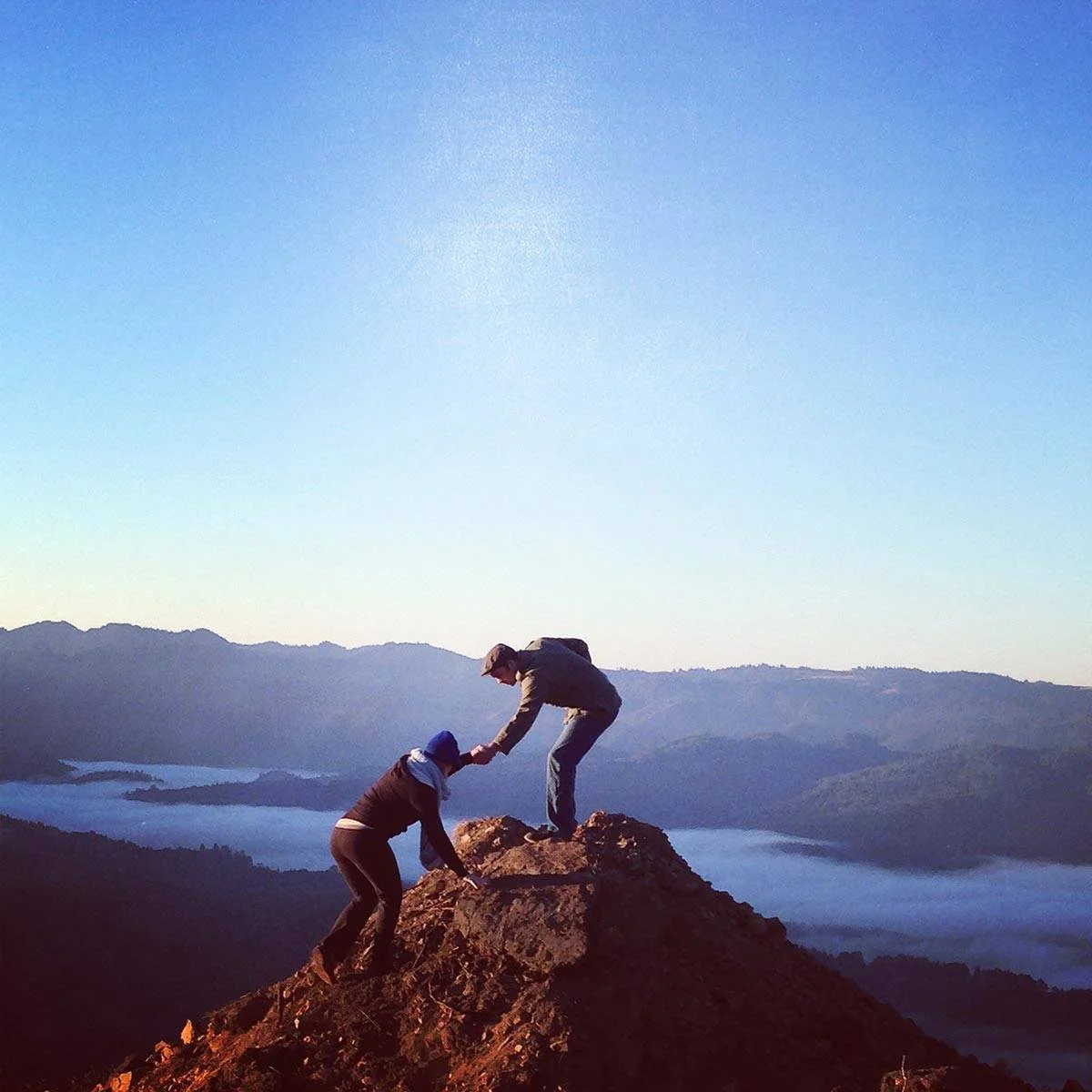 Two people on a mountain summit, one helping the other to climb up, with a scenic landscape of mountains and a lake in the background under a clear blue sky.