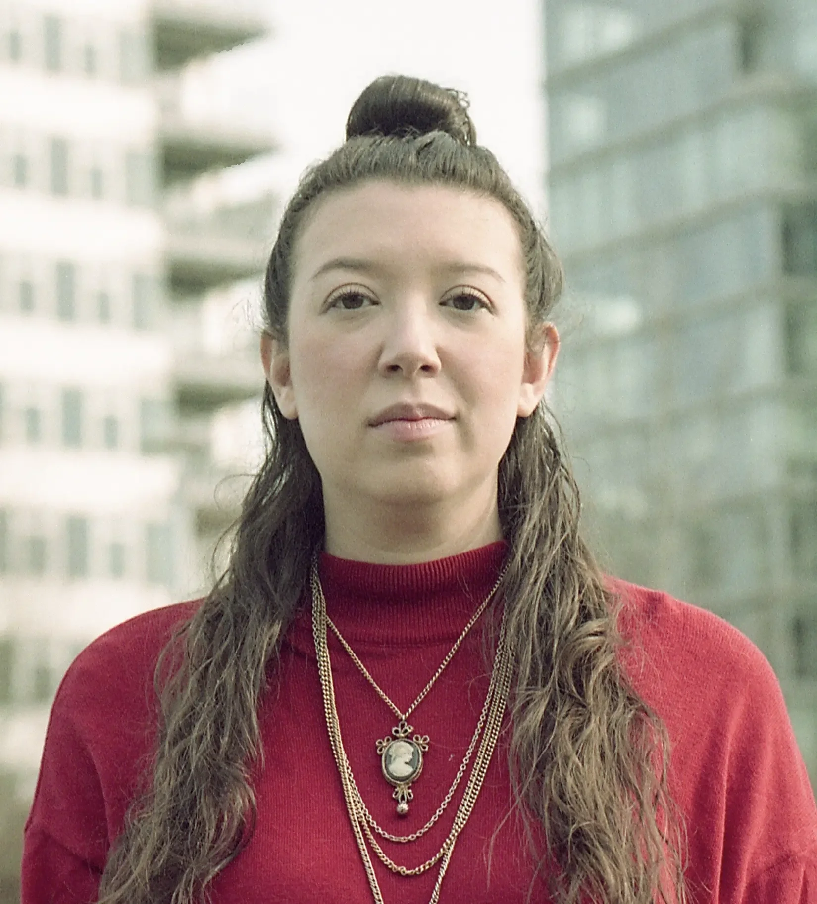 A woman with long wavy brown hair tied in a bun, wearing a red sweater and layered gold necklaces, standing outdoors with a blurred building in the background.