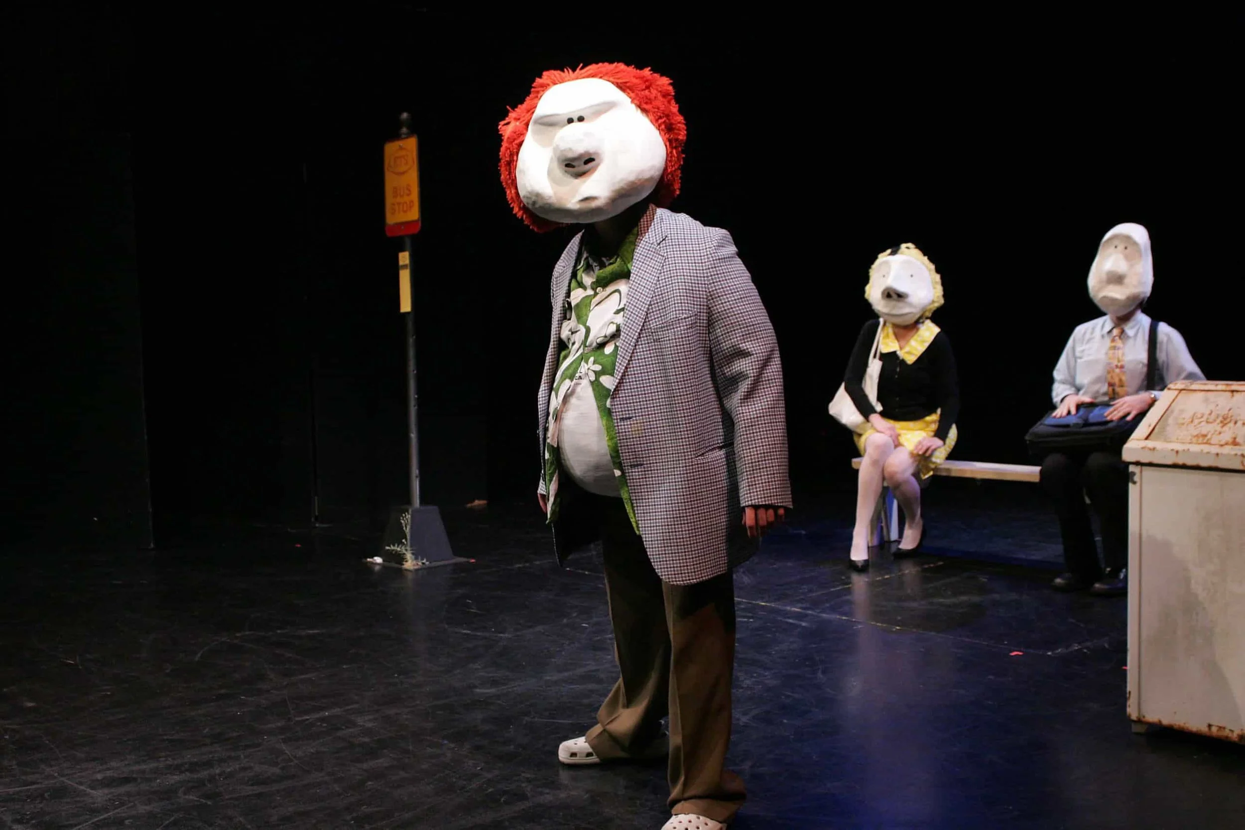 Person wearing a lion mask, a blazer, and brown pants standing on a stage with two seated individuals in masks in the background.