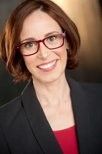 A woman with short brown hair, wearing red glasses, smiling, dressed in a black blazer over a red top.