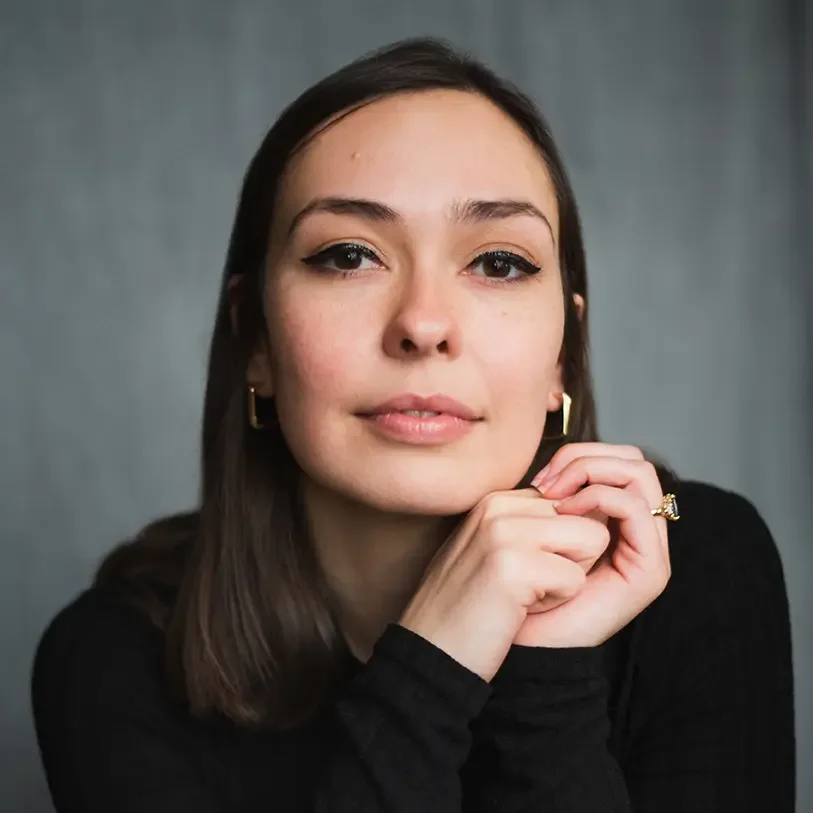 A young woman with brown hair, wearing a black top, gold earrings, and a gold ring, resting her chin on her hands, looking at the camera with a neutral expression.