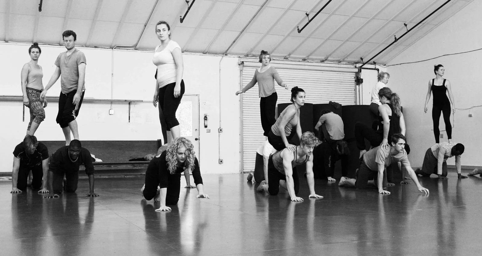 Group of people practicing yoga or posing in a dance studio with a high ceiling, some balancing on their hands and knees, with others standing or crouching.