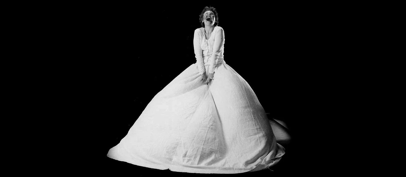 A woman in a long white dress sits on a large voluminous skirt, laughing with her head tilted back, in a black and white photo with a plain dark background.