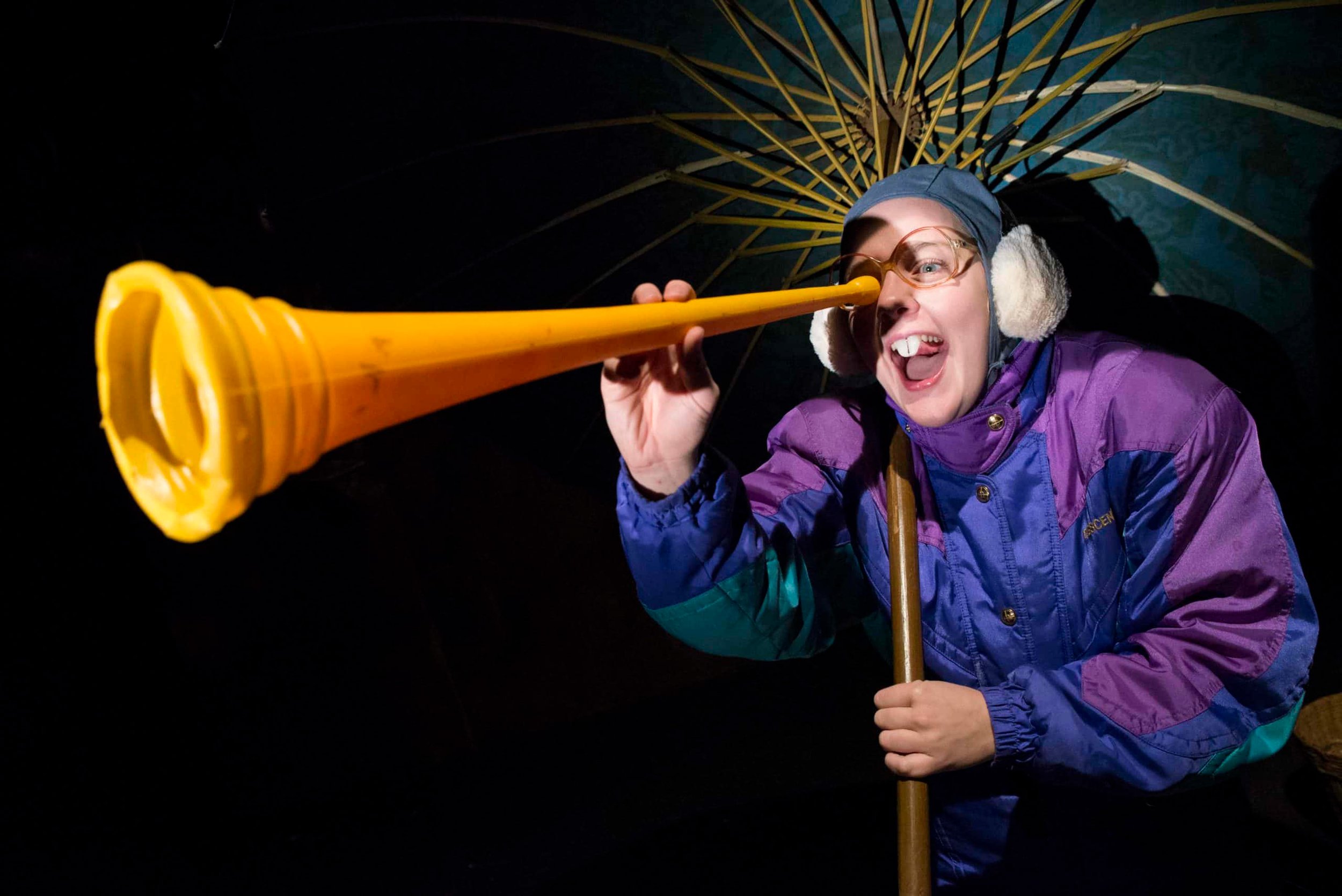 Person in outdoor gear holding a yellow alpine horn with an open mouth, wearing glasses, a blue hat, and ear protection, standing under a large yellow umbrella at night.