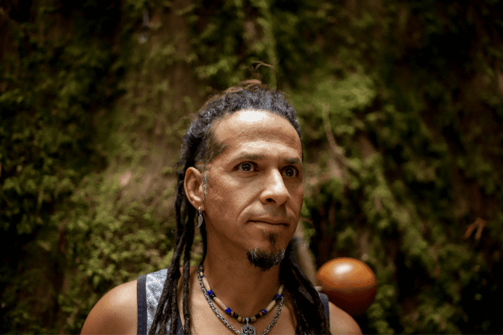 A man with long dreadlocks, wearing earrings, necklaces, and a sleeveless shirt, standing outdoors with green foliage and a brown ceramic pot in the background.