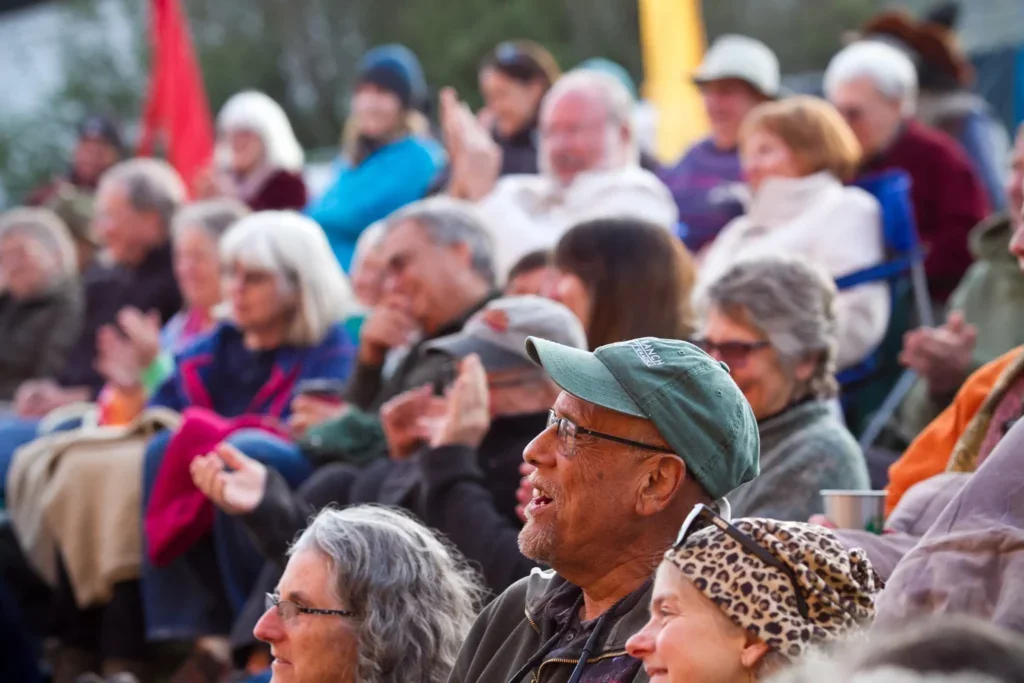 A diverse group of elderly people sitting outdoors, enjoying a performance or event, with some clapping and smiling.