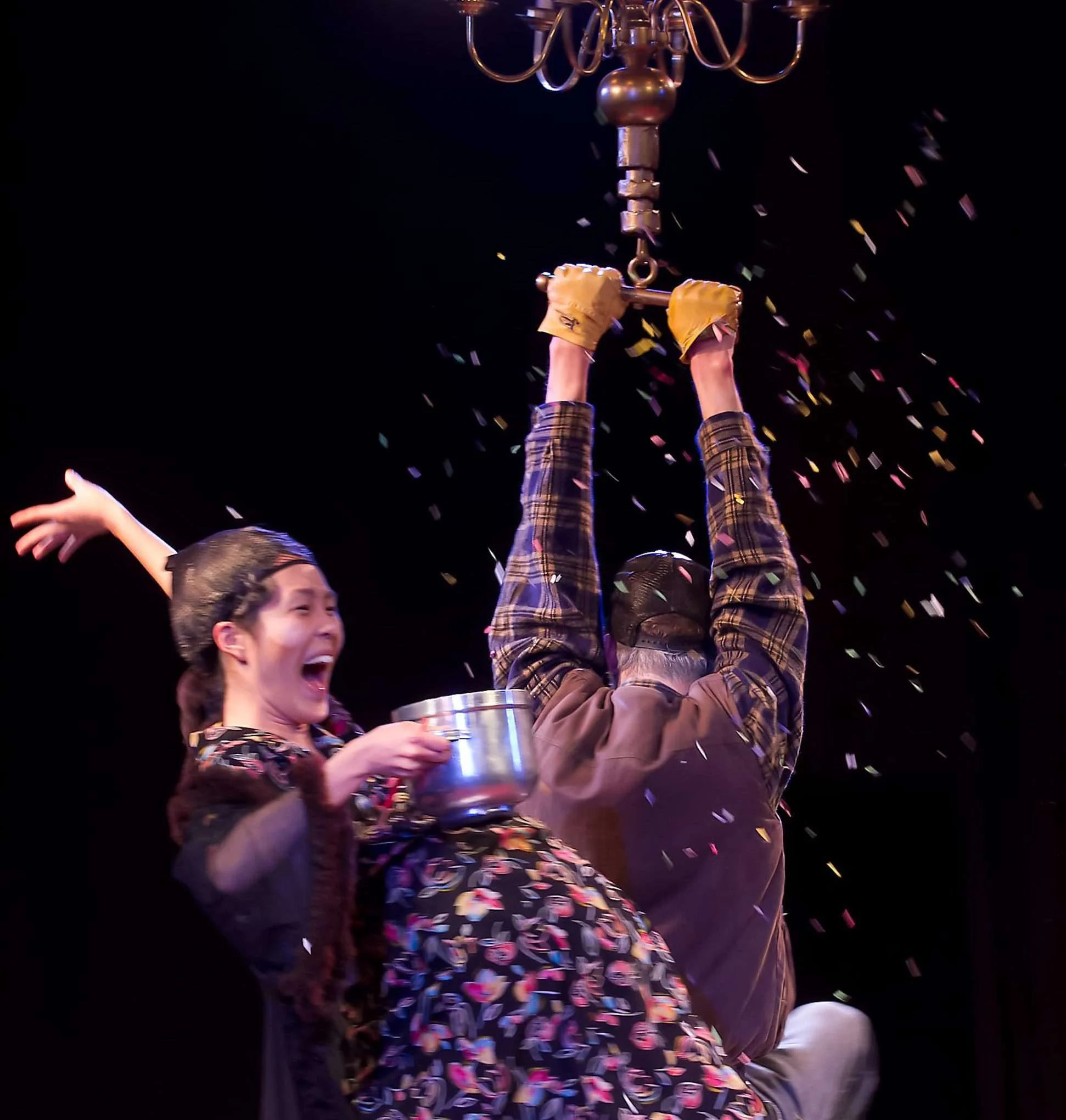 People celebrating with confetti, holding a trophy, on stage with a chandelier overhead.