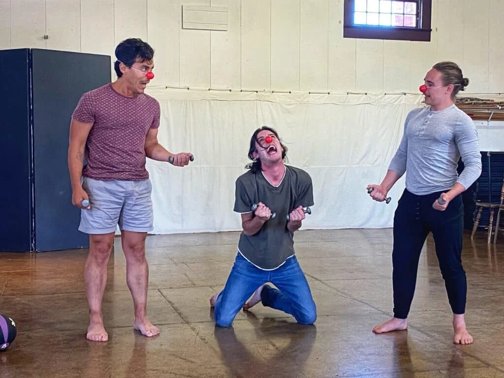 Three people dressed as clowns with red noses, holding small mallets, standing and kneeling in a room with a wooden floor and white walls, appearing to be laughing or joking.