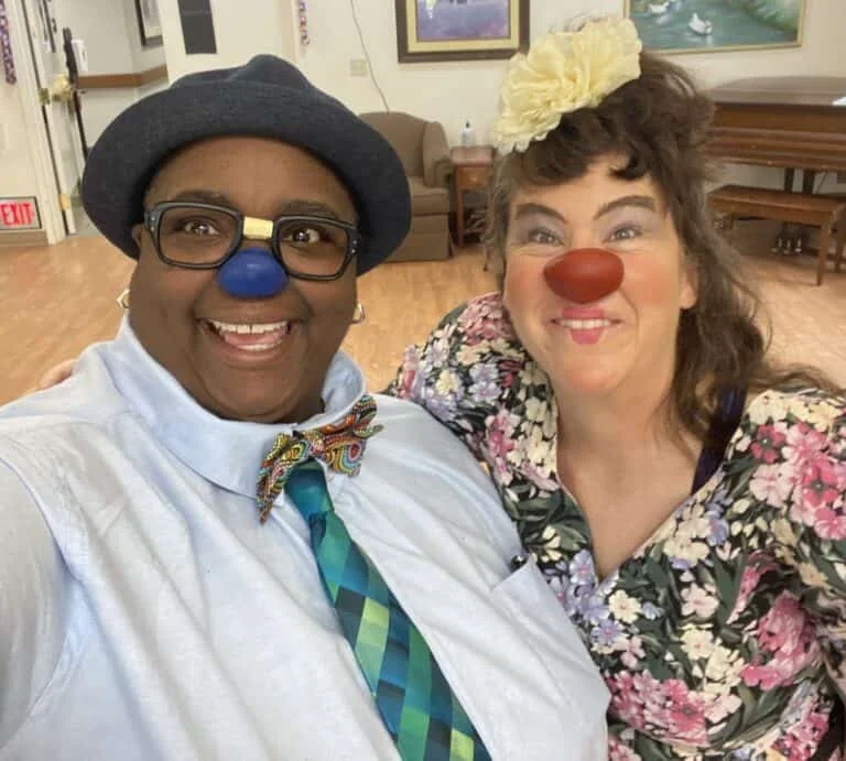 Two people dressed as clowns taking a selfie, both with red noses and cheerful expressions, in an indoor setting with wooden floors and art on the walls.
