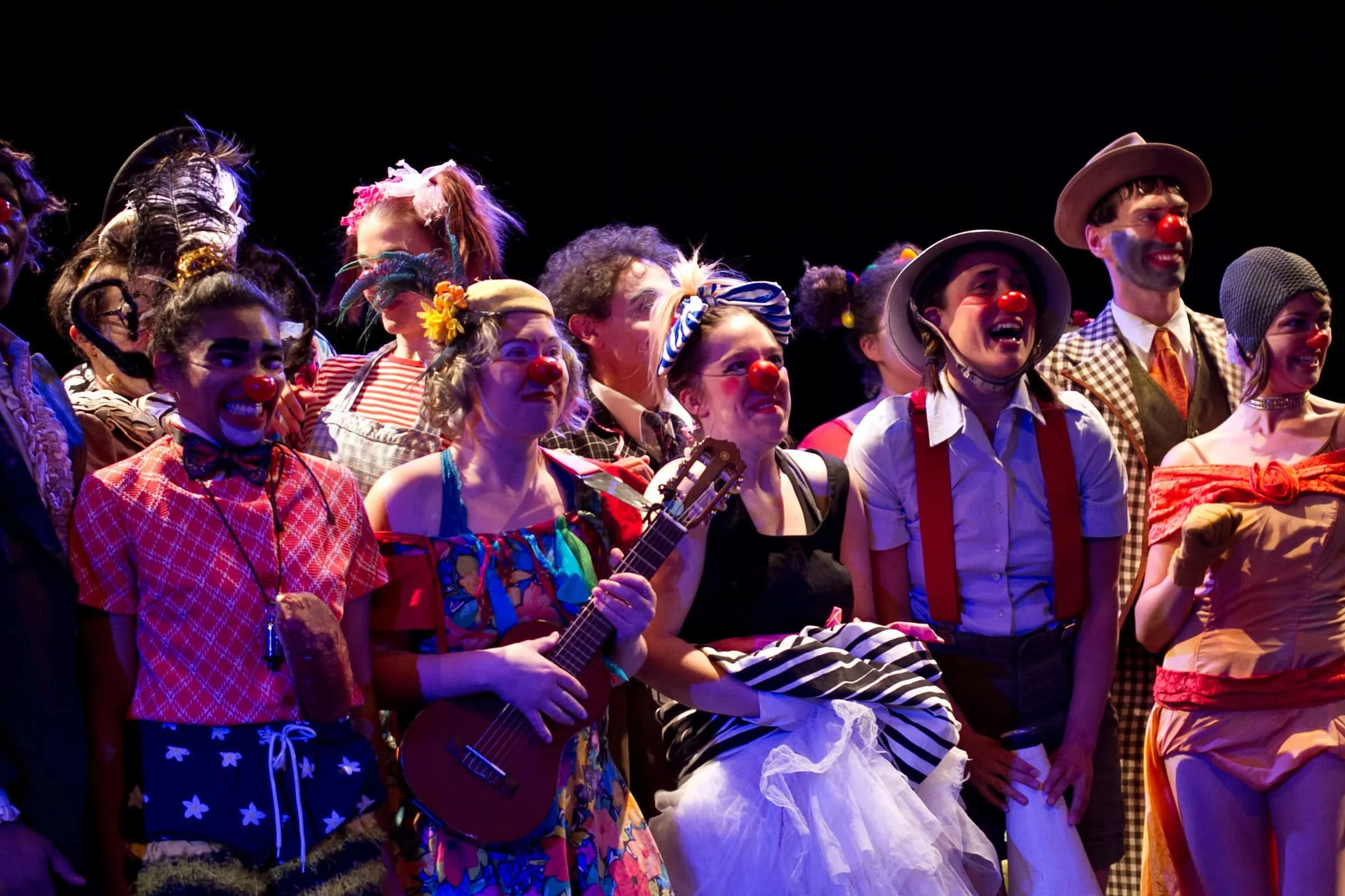 A group of performers dressed as clowns and circus characters on stage, wearing colorful costumes, makeup, hats, and red noses.