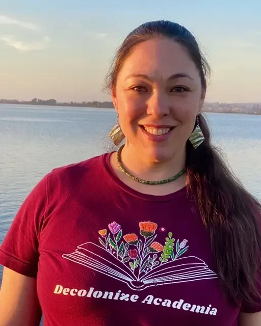 A young woman with long dark hair, wearing a maroon t-shirt with an open book and flowers design, standing near a body of water during sunset.