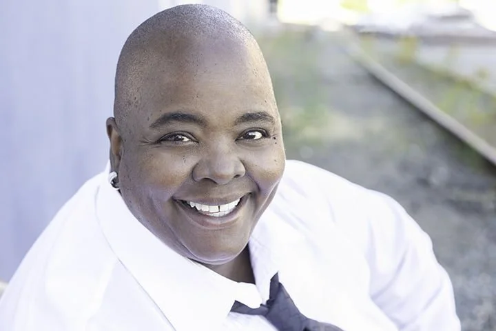 Close-up of a smiling African American woman with a shaved head, wearing a white shirt and a loosened black tie, outdoors with train tracks in the background.